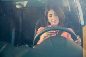 A younger woman driving in a car, focused on her phone, which she holds at steering wheel level