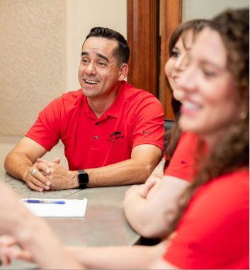 Un hombre con una camisa de polo roja se sienta en una mesa de conferencias de About Us junto a dos colegas, sonriendo mientras mira hacia alguien que no está en la cámara.