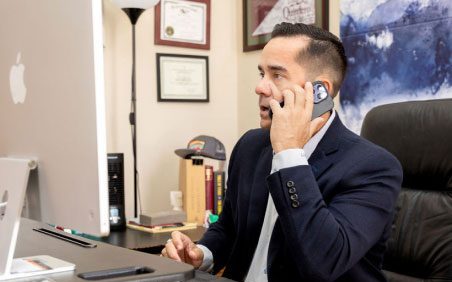 Un hombre con una chaqueta de traje hablando por el teléfono inteligente mientras se sienta en la mesa de Atención al Cliente con una computadora.