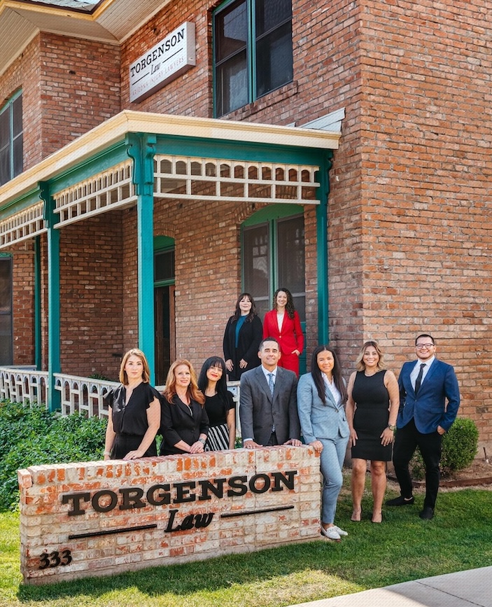 A group of eight people stand in front of a brick building with a sign that reads Torgenson Law, with a matching stone sign in the foreground.