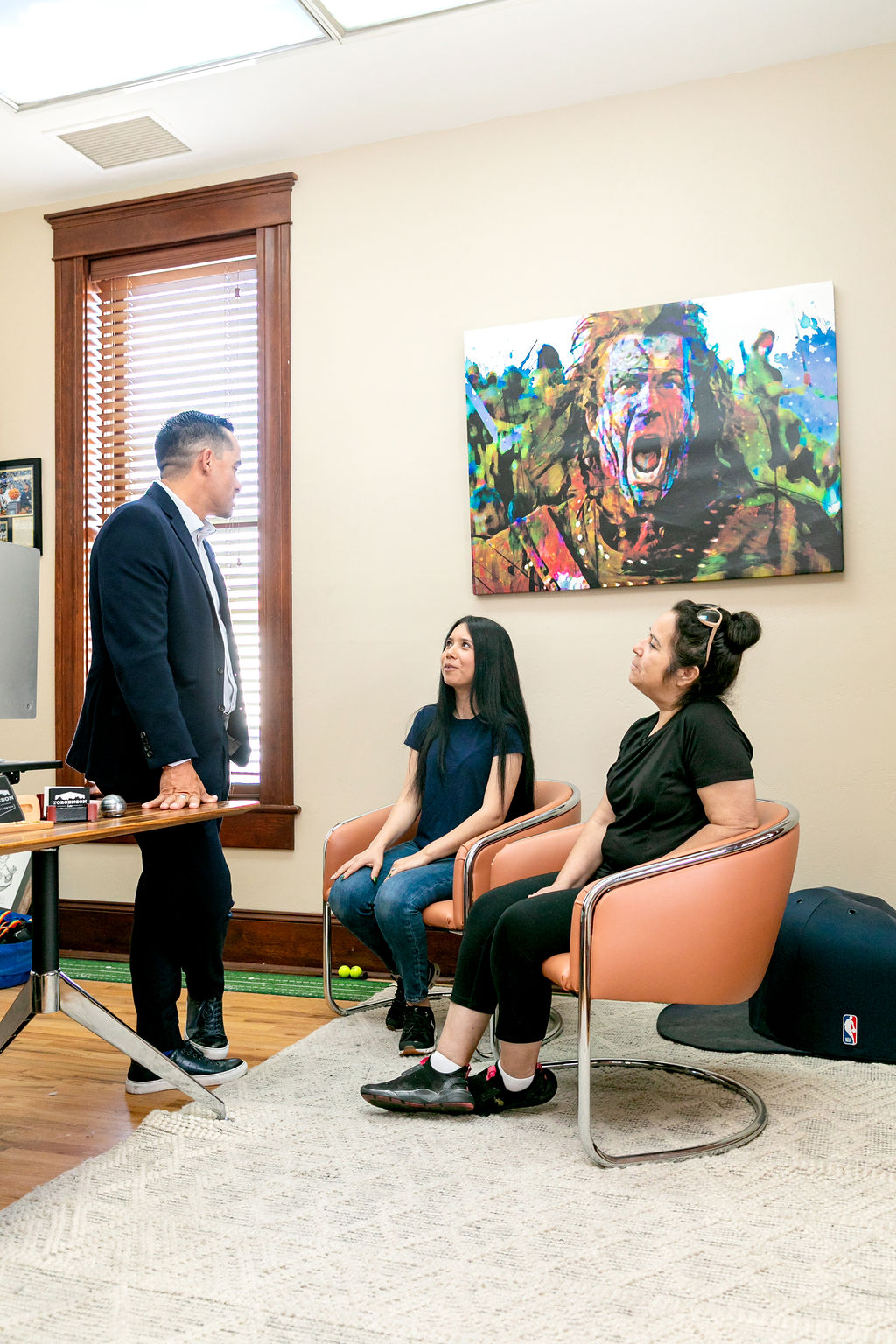 A man in a suit stands and speaks to two women seated in chairs, beneath a large painting of a screaming man.