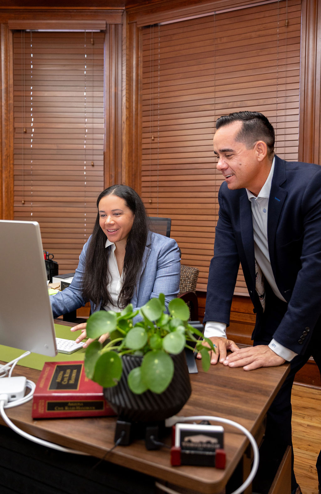 A man in a suit leans over a desk to look at a computer screen with a smiling woman in a blazer.
