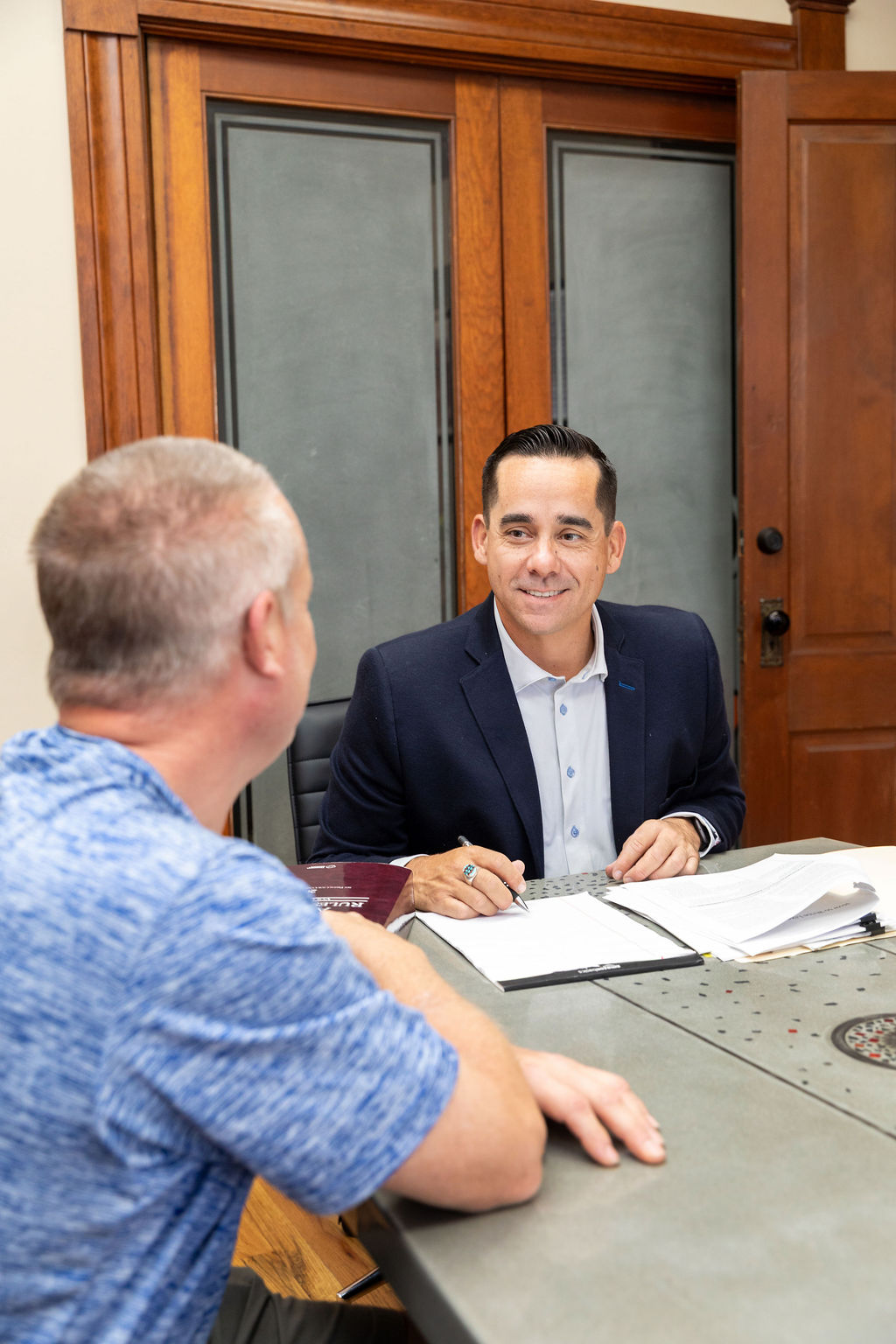 A man in a suit jacket signs a document while sitting across a table from another man in an office.