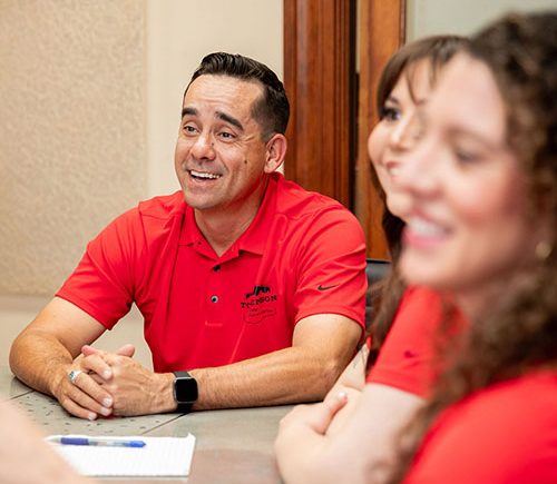 A smiling man in a red polo shirt sits at a table with two colleagues.