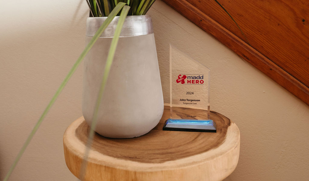 Un trofeo de premio MADD Hero 2024 para John Torgenson de Torgenson Law sentado en una mesa de madera junto a una planta en maceta, como se muestra en nuestras preguntas frecuentes.