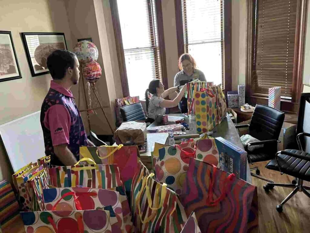 Three people assemble Christmas gift bags at a table covered in colorful, patterned bags and boxes in a sunlit office.