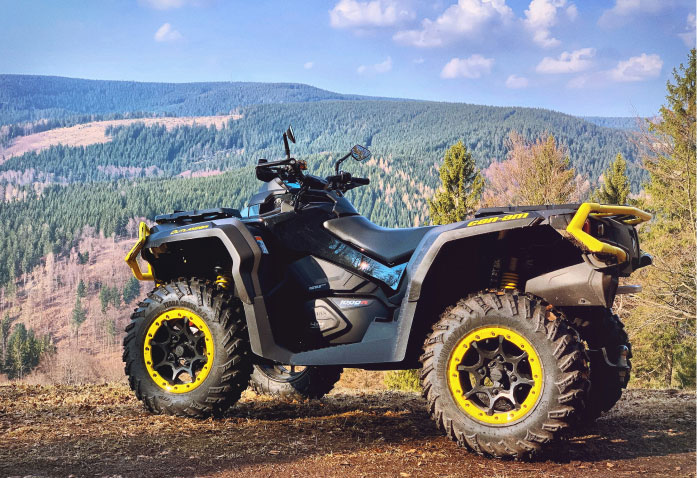 A black Can-Am Outlander ATV with yellow accents parked on a dirt trail that could pose a personal injury risk, overlooking a forested mountain range.