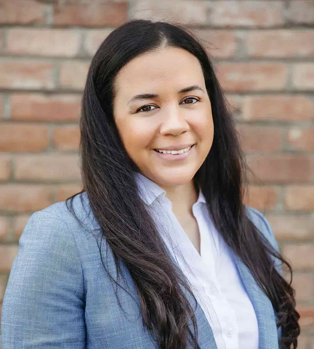 A professional headshot of Ariana Byrd, smiling and wearing a white collared shirt and a blue blazer in front of a brick wall.