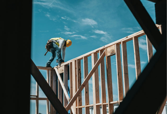 A construction worker wearing a yellow hard hat and safety vest stands on a wooden frame wall, representing a potential personal injury risk while working.