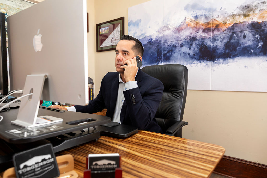 A man in a suit jacket sits at a desk, speaking on a smartphone while looking at a computer monitor.