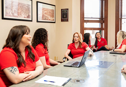 A group of women in matching red polo shirts sit around a conference table during a meeting discussing the Fair Fee Guarantee.
