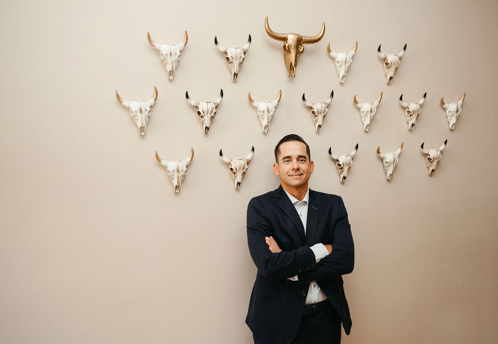 A professional portrait of John Torgenson in a dark navy suit and white shirt, standing with his arms crossed in front of a beige wall decorated with several white animal skulls arranged in a symmetrical pattern.