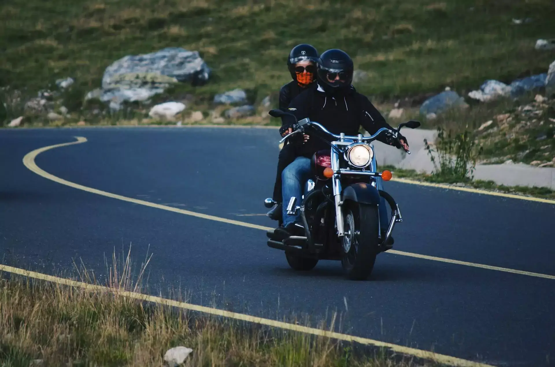 Two people in helmets ride a motorcycle along a curved mountain road that is the site of a motorcycle accident.