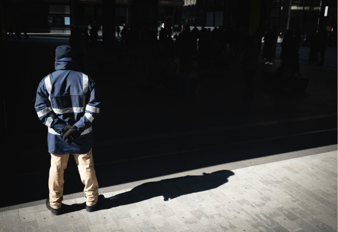 A security guard in a dark blue jacket and tan pants stands with hands behind his back, casting a long shadow on a paved surface often associated with personal injury risks.