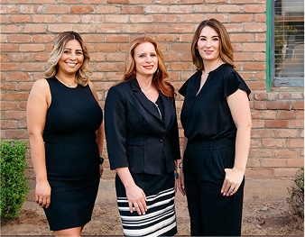 Tres mujeres profesionales que se encuentran una al lado de la otra frente a una pared de ladrillo, listas para responder a las preguntas más frecuentes.