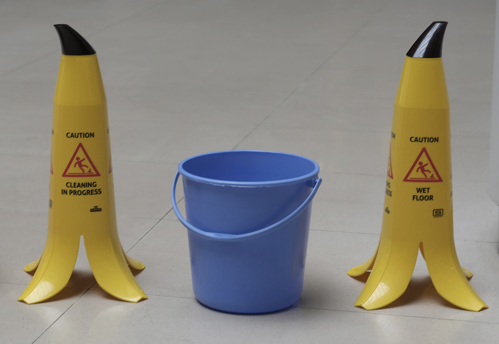 Two yellow, banana-shaped slip and fall caution cones standing on either side of a blue bucket.