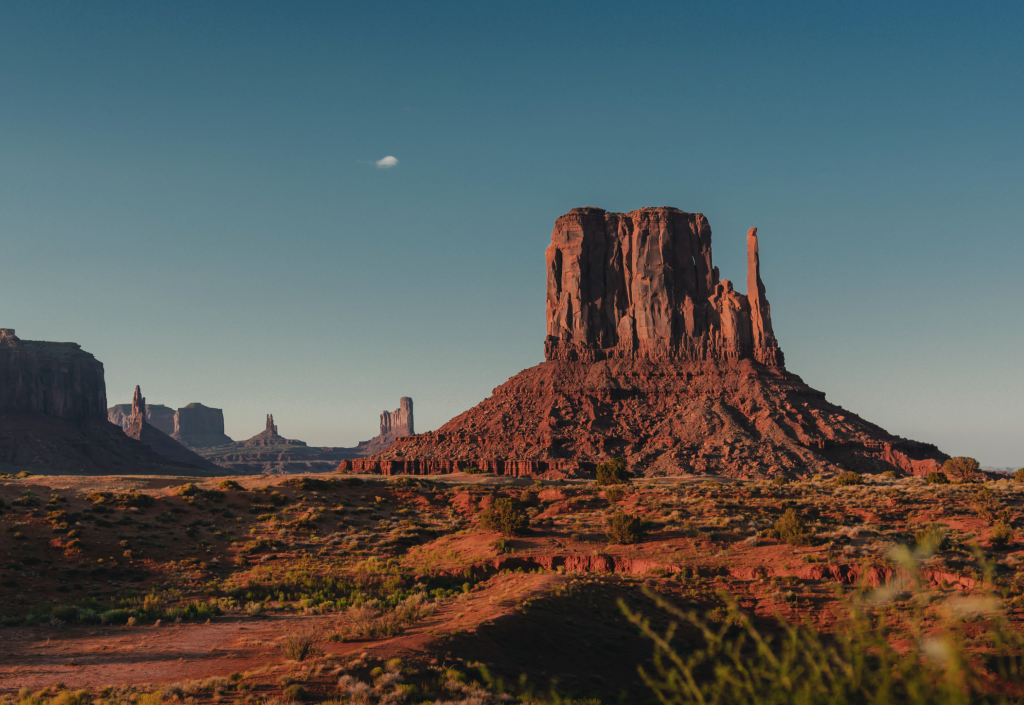 The West Mitten Butte rises above the desert floor and its ancient fault lines in Monument Valley under a clear blue sky.