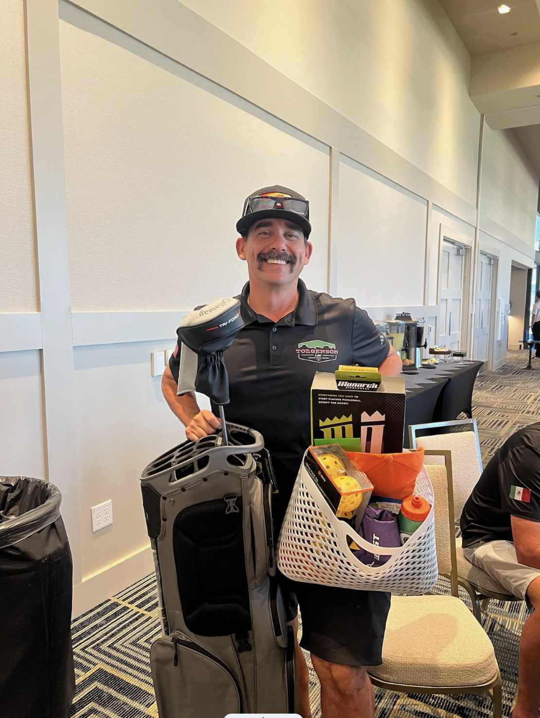 A smiling man, embodying a Latina Strong spirit, wears a Tolgerson Lake polo shirt and cap while holding a golf bag in one hand and a basket of pickleball equipment in the other.