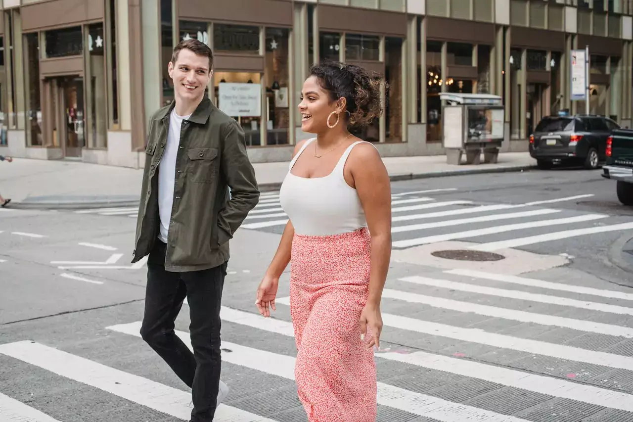 A man in a green jacket and a woman in a white tank top and floral skirt walk across a city crosswalk.