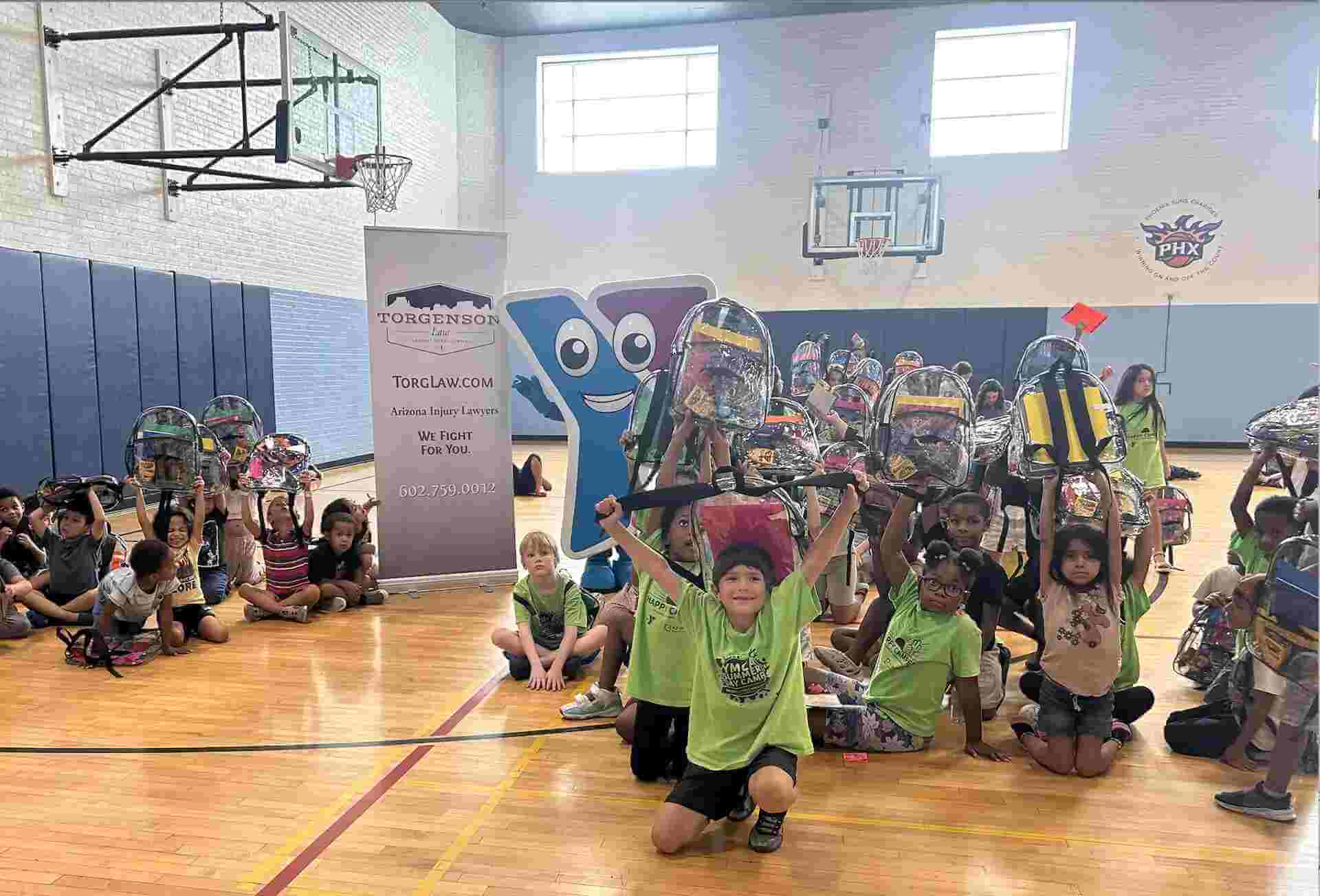 Children hold up clear backpacks during a YMCA backpack drive in a gymnasium next to a Torgenson Law banner and a YMCA mascot.