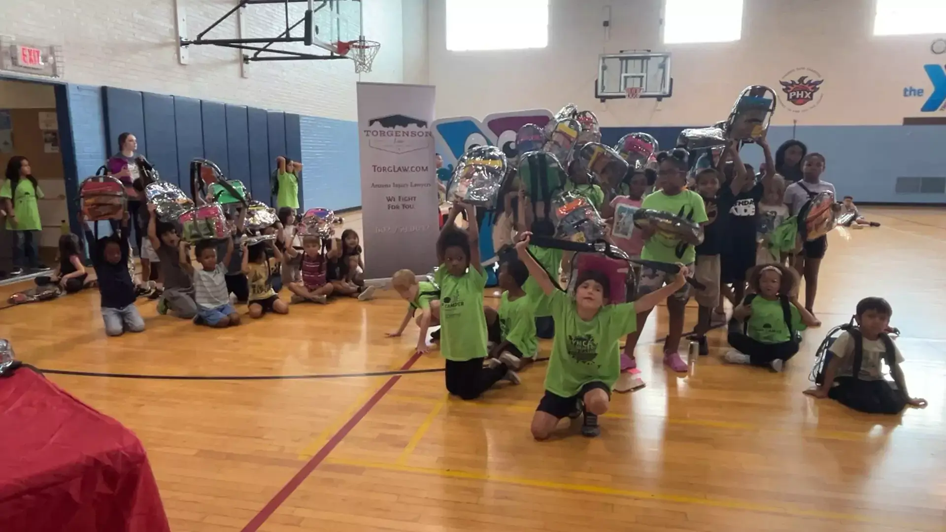 A large group of children in a gymnasium hold up backpacks above their heads during a Backpack Drive in front of a Torgenson law firm banner.
