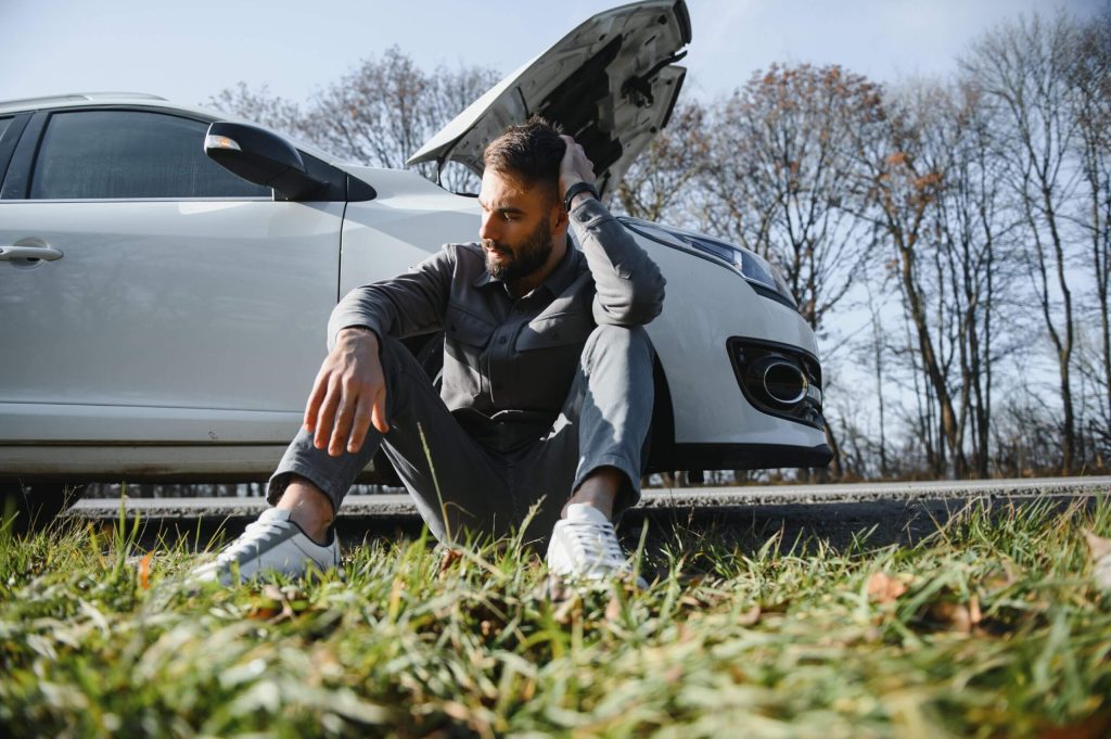Man sitting on grass by a white car with an open hood with his hand on his head