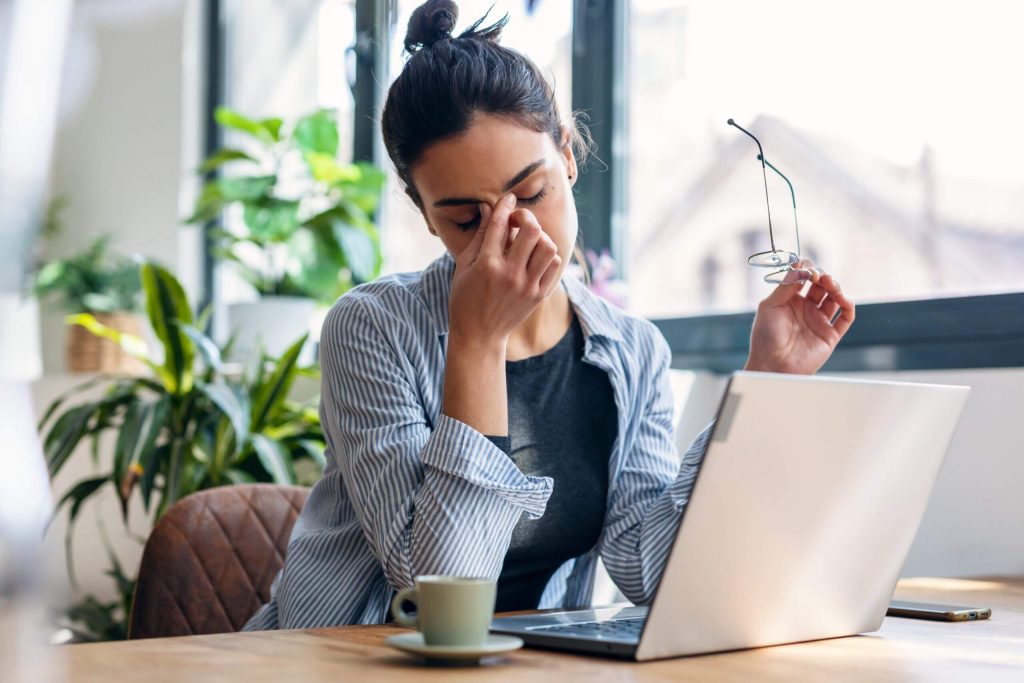 A woman sits at a desk with a laptop, holding her head from a headache.