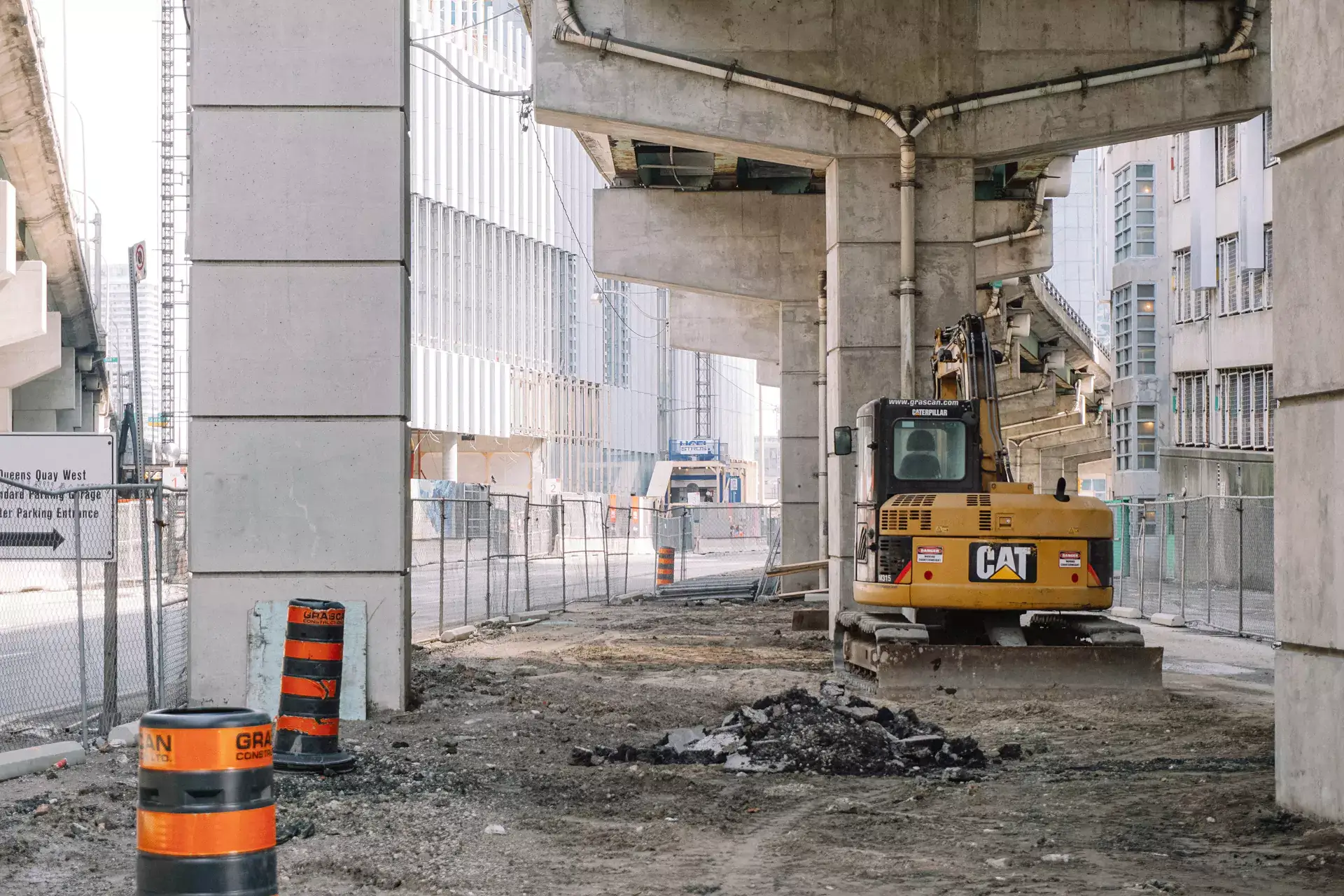 A yellow CAT excavator parked on a dirt lot beneath a concrete overpass near an ATV, with construction barrels in the foreground and a fenced construction site in the background.
