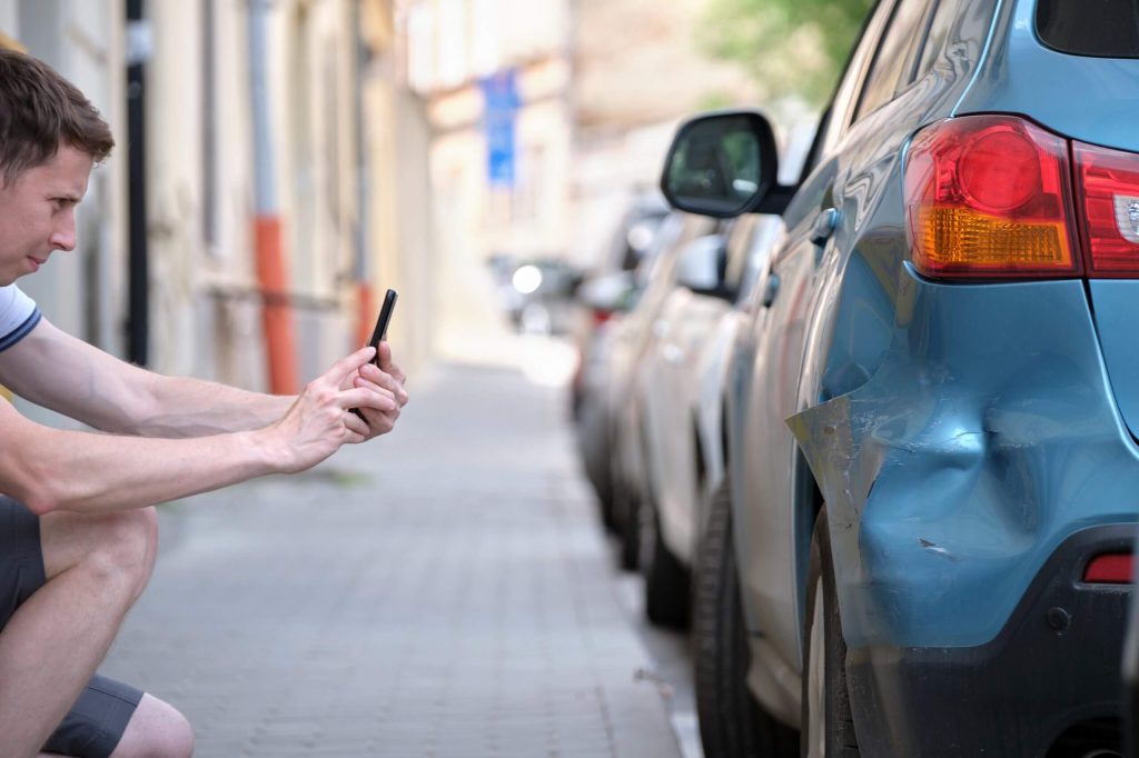 A person kneels on a sidewalk, photographing a blue car's dented rear bumper with a smartphone