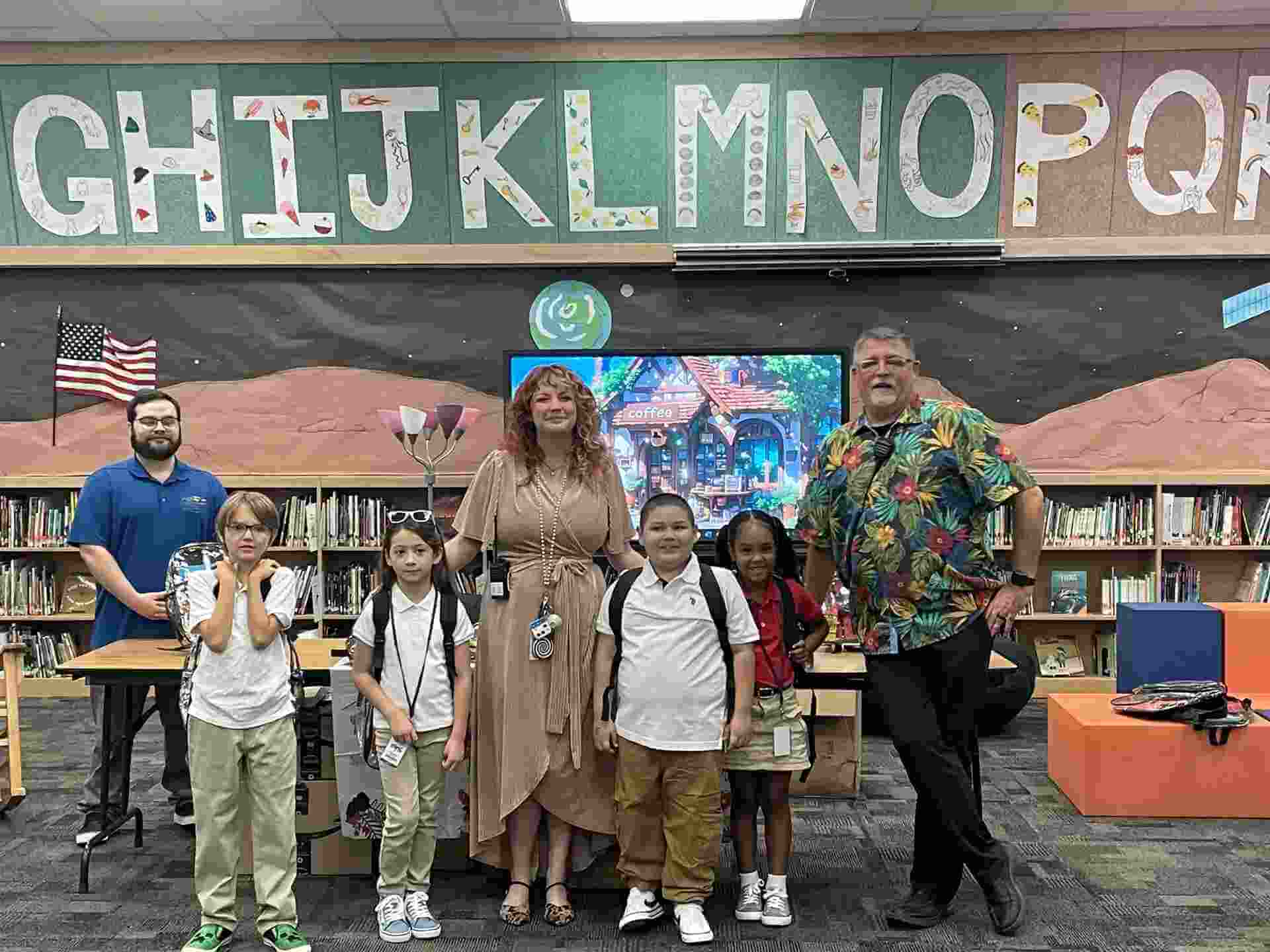 Two adults and four children stand in a school library for a backpack drive beneath a wall decorated with large letters of the alphabet.