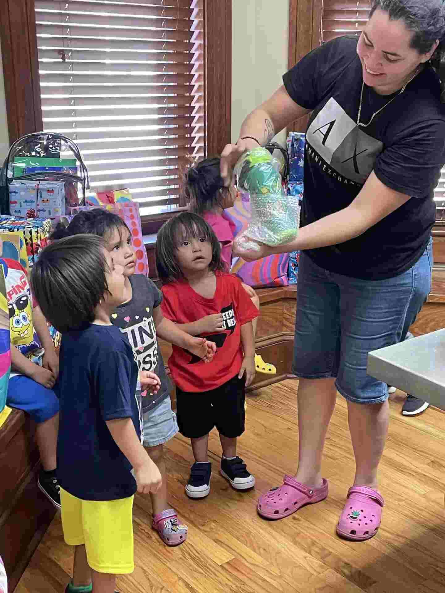 A woman hands a small, bubble-wrapped Christmas gift to a group of young children.