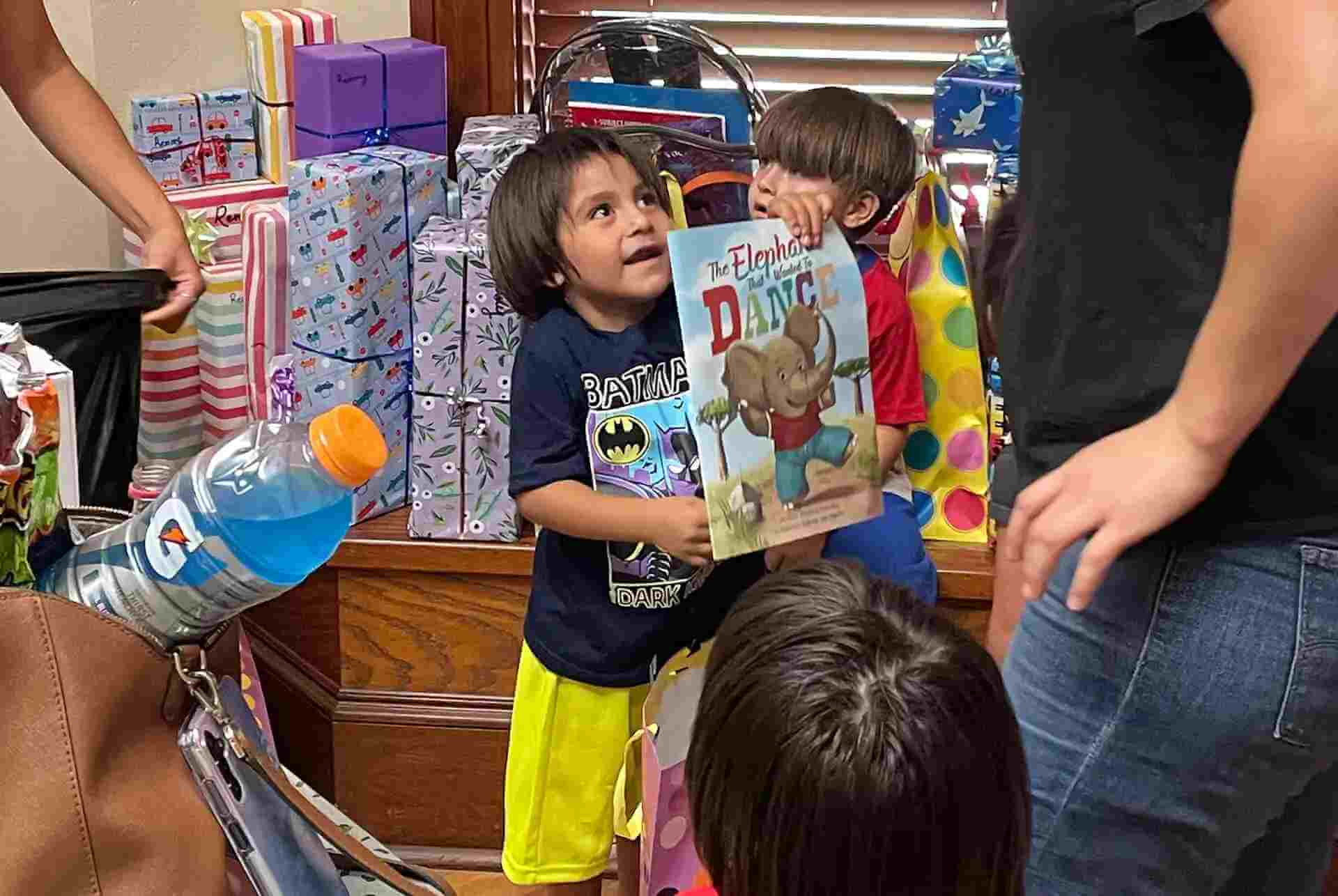 A young child holds a copy of The Elephant Who Wanted To Dance while standing among stacks of wrapped Christmas gifts.