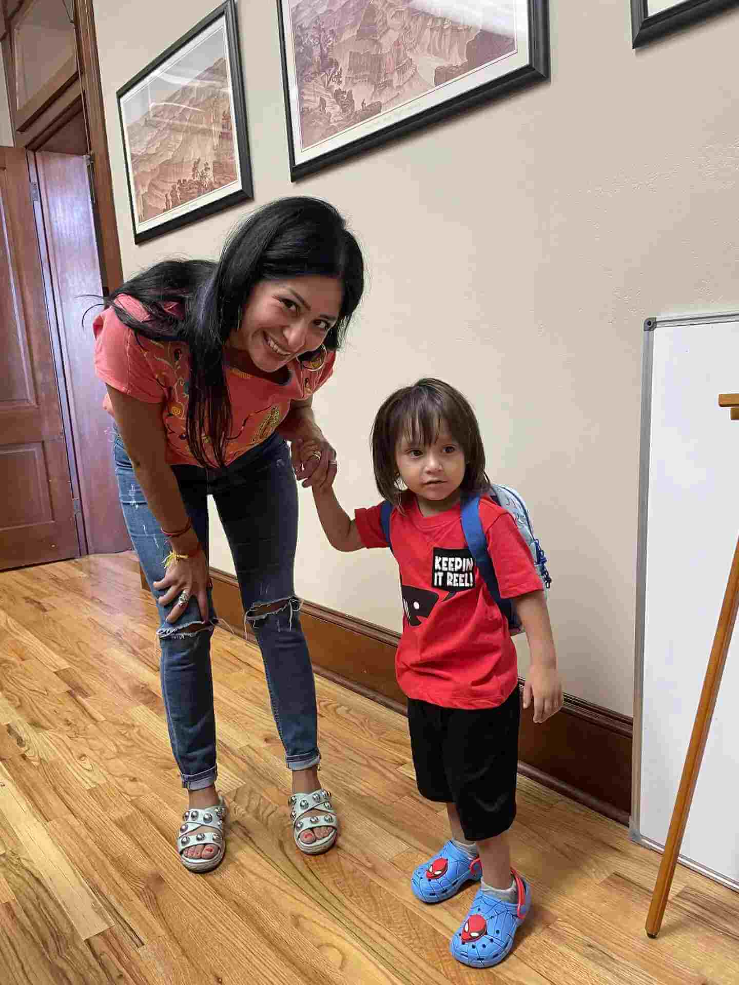 A woman standing in a Christmas hallway holds the hand of a young child wearing a red t-shirt that says Keepin It Reel.