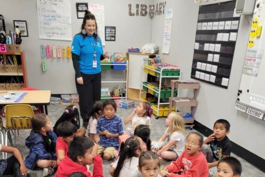 A teacher stands in front of a classroom of young students sitting on the floor for a lesson.