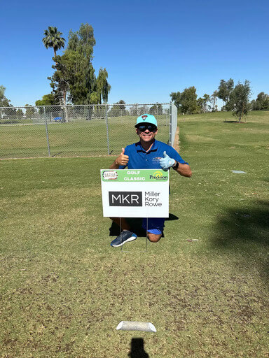 A man in a blue polo shirt and hat kneels on a golf course holding a sign for MAM’s 11th Annual Golf Classic that reads Golf Classic and Miller Kory Rowe.