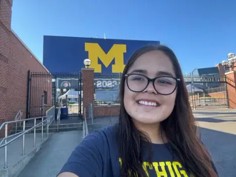 A smiling young woman in a Michigan t-shirt takes a selfie in front of the University of Michigan stadium entrance.