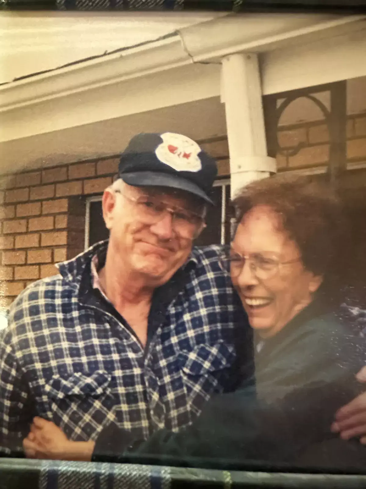 An elderly man in a plaid shirt and baseball cap smiles while being hugged by an elderly woman wearing glasses, captured at the 2024 Renee Torgenson Scholar event.