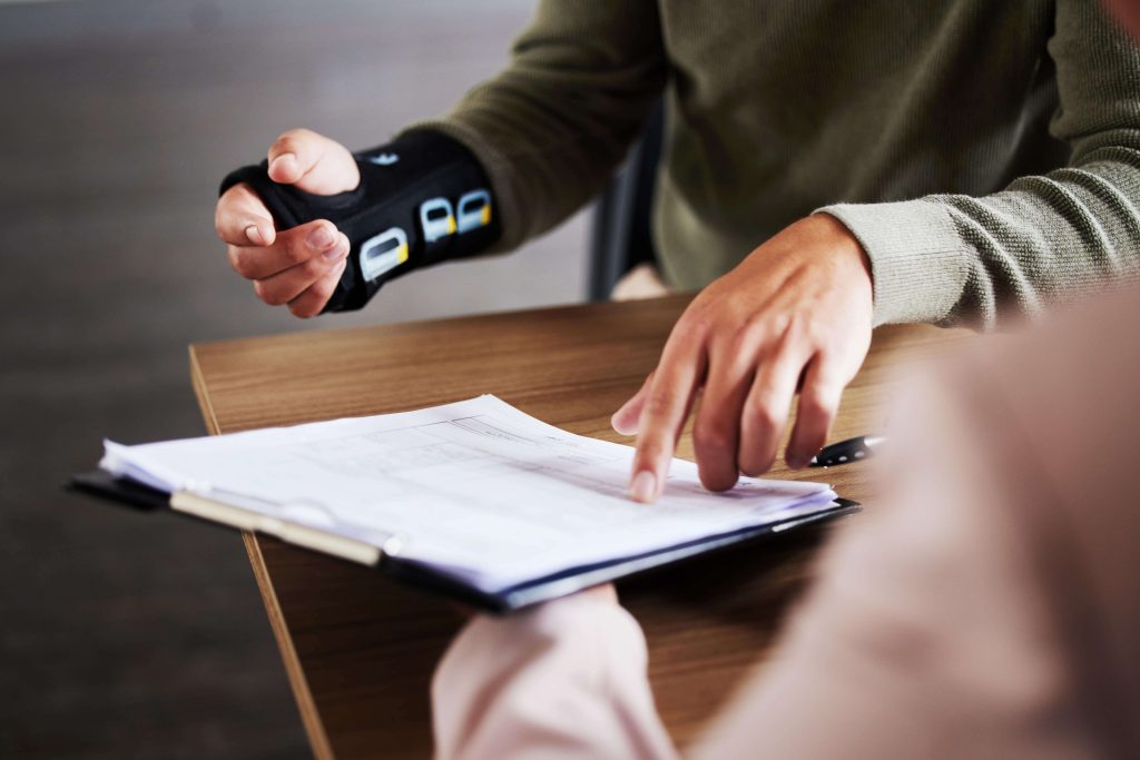 A person with a wrist support gestures while another points at a document on a clipboard]
