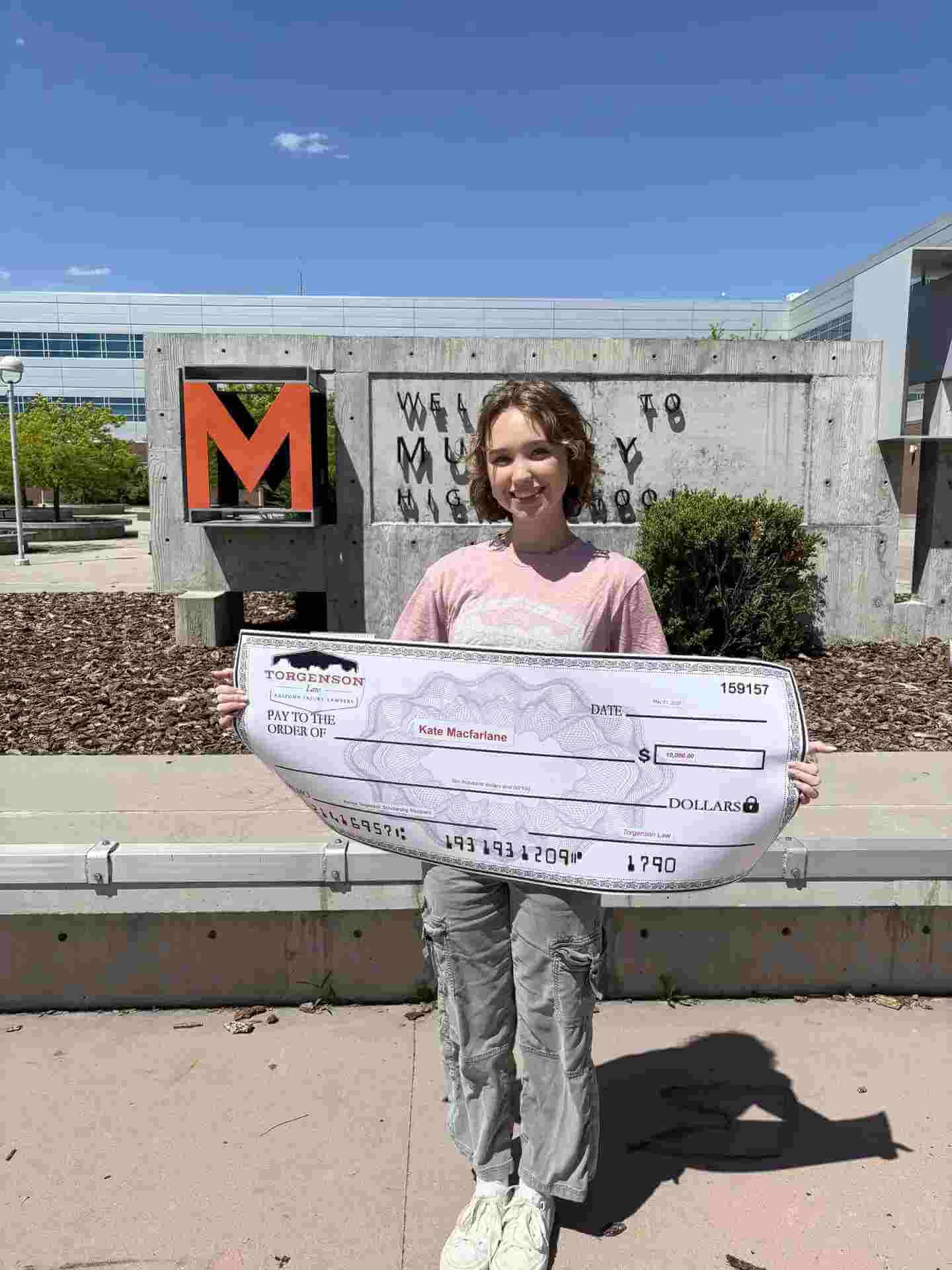 Renee Torgenson stands in front of a concrete monument holding a large novelty check made out to Kate Macfarlane for $10,000.