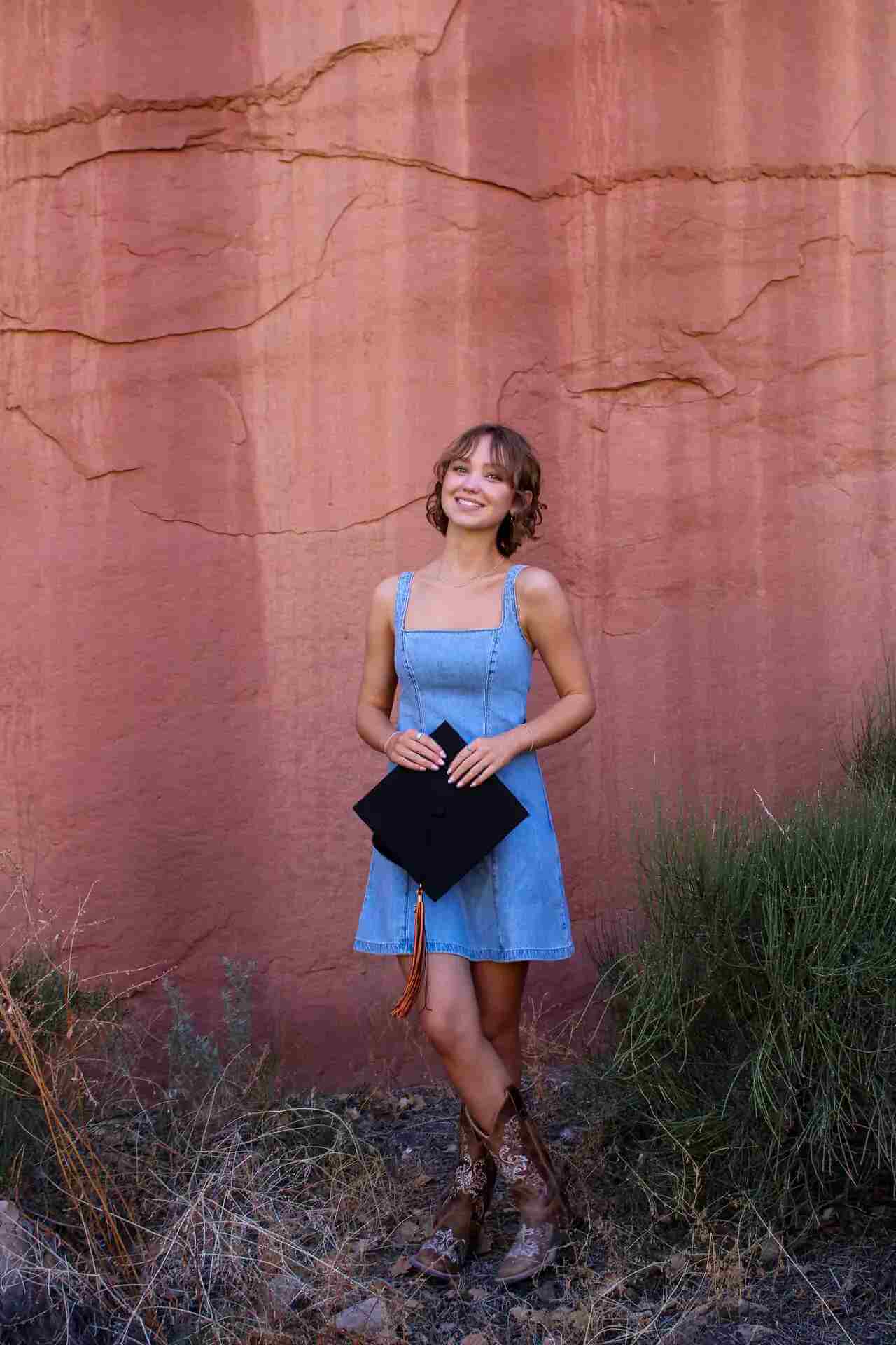 Renee Torgenson smiles while wearing a denim dress and cowboy boots, holding a graduation cap in front of a red rock wall.