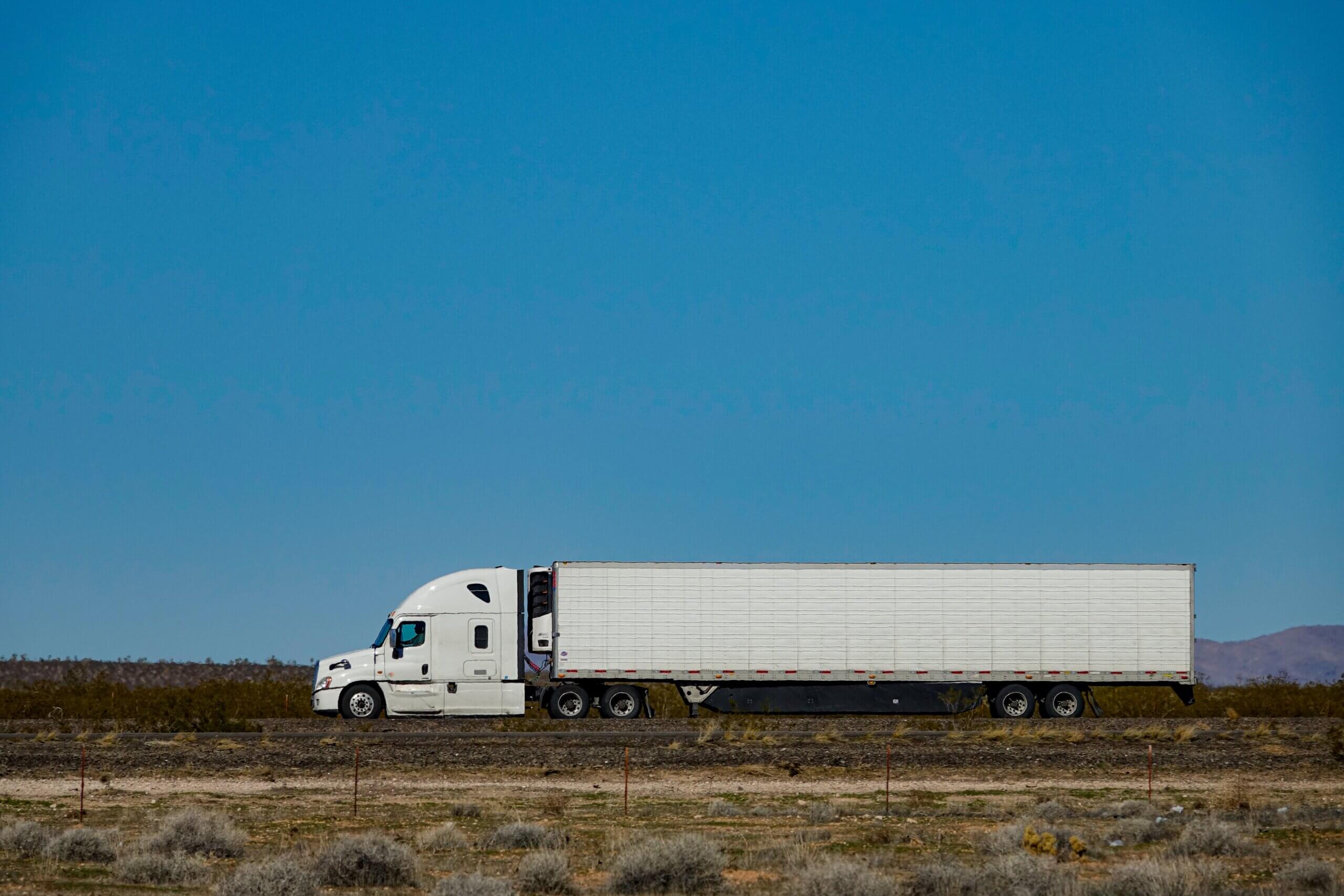 A white semi-truck travels along a desert highway under a clear blue sky, evoking a sense of long-haul fatigue.