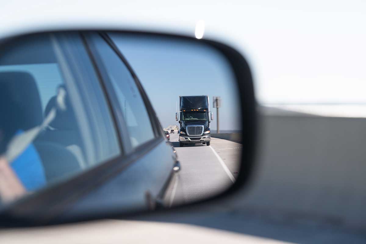 A semi-truck looms in a car’s side-view mirror while driving on a bridge, evoking a sense of road rage.
