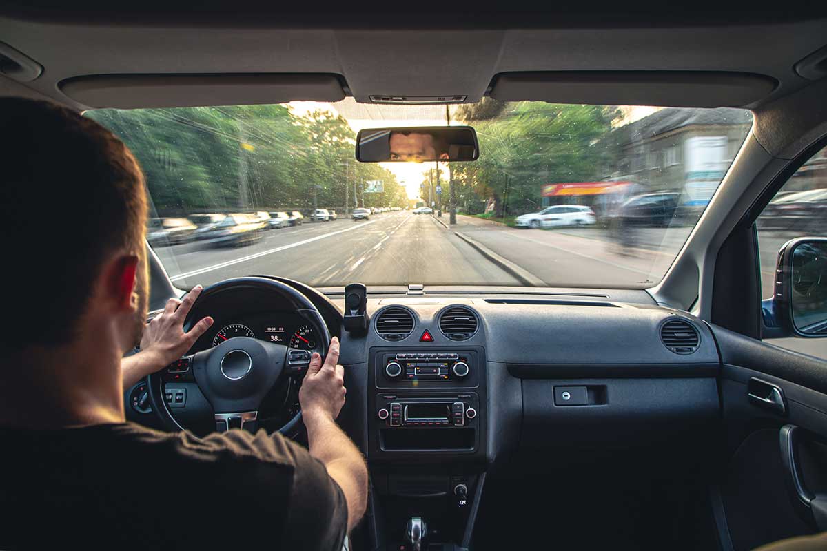 Una vista desde el asiento trasero de un coche, que muestra al conductor detrás del volante mientras recorre una carretera iluminada por el sol.
