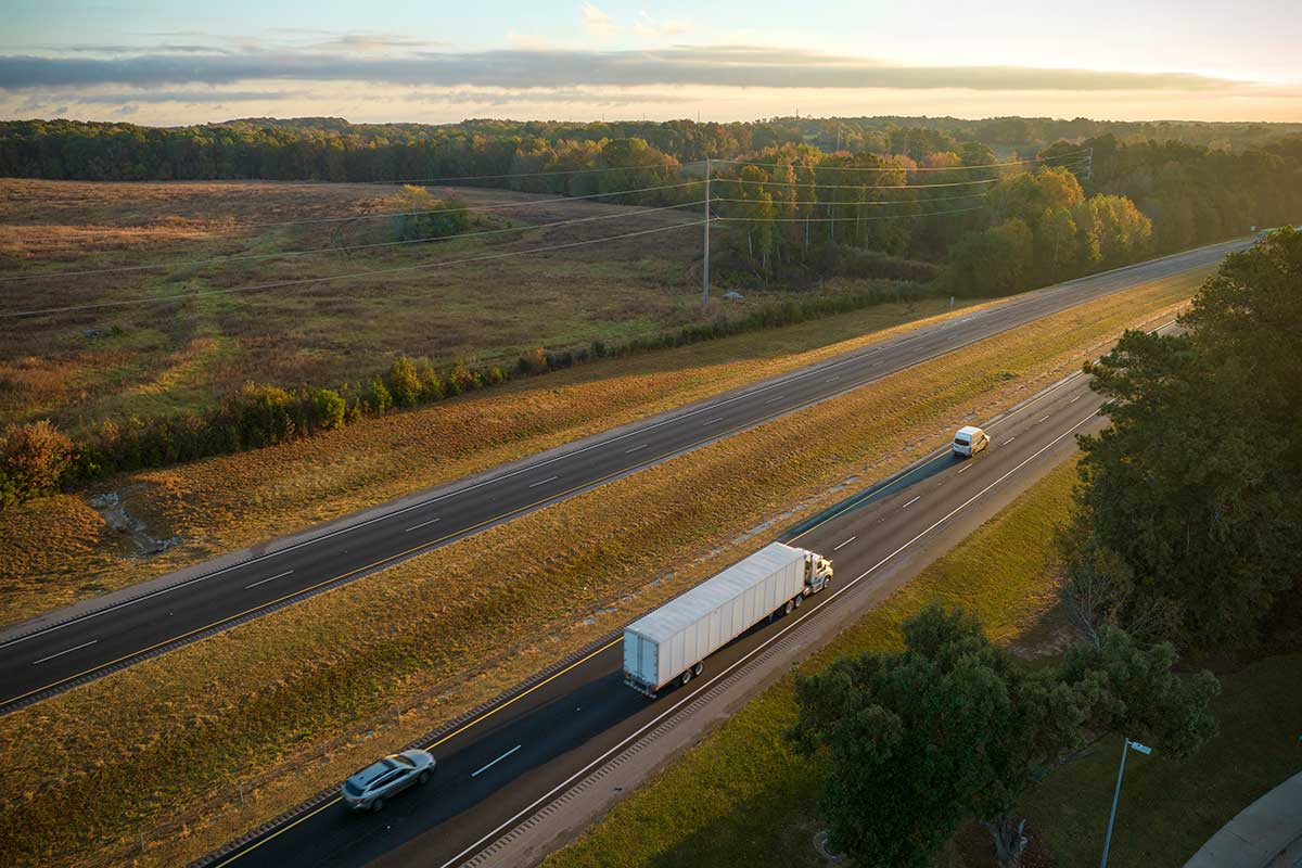 A semi-truck and two cars traveling on a highway through a rural landscape at sunrise.