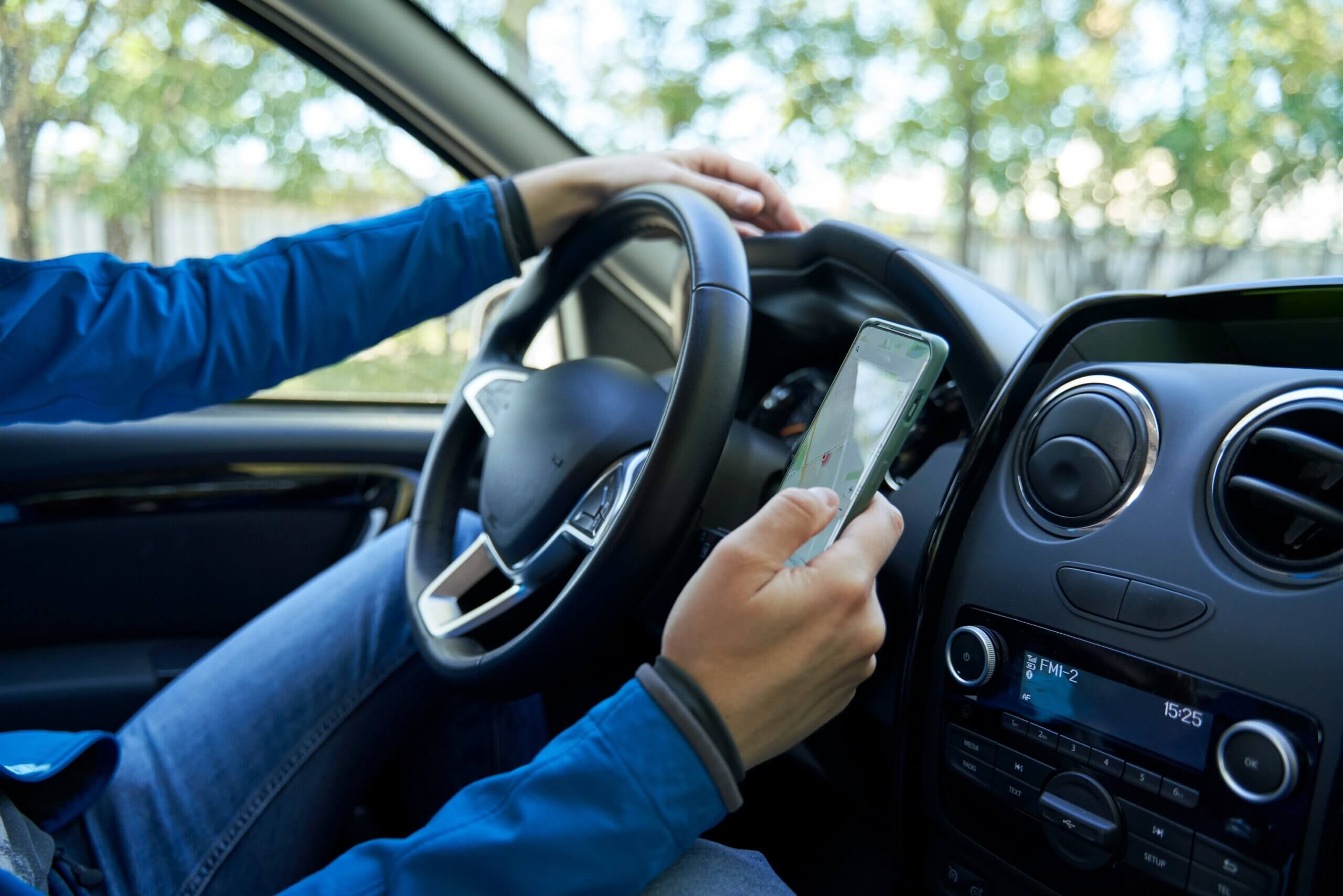 A rideshare driver in a blue jacket driving a car while holding a smartphone displaying a map.