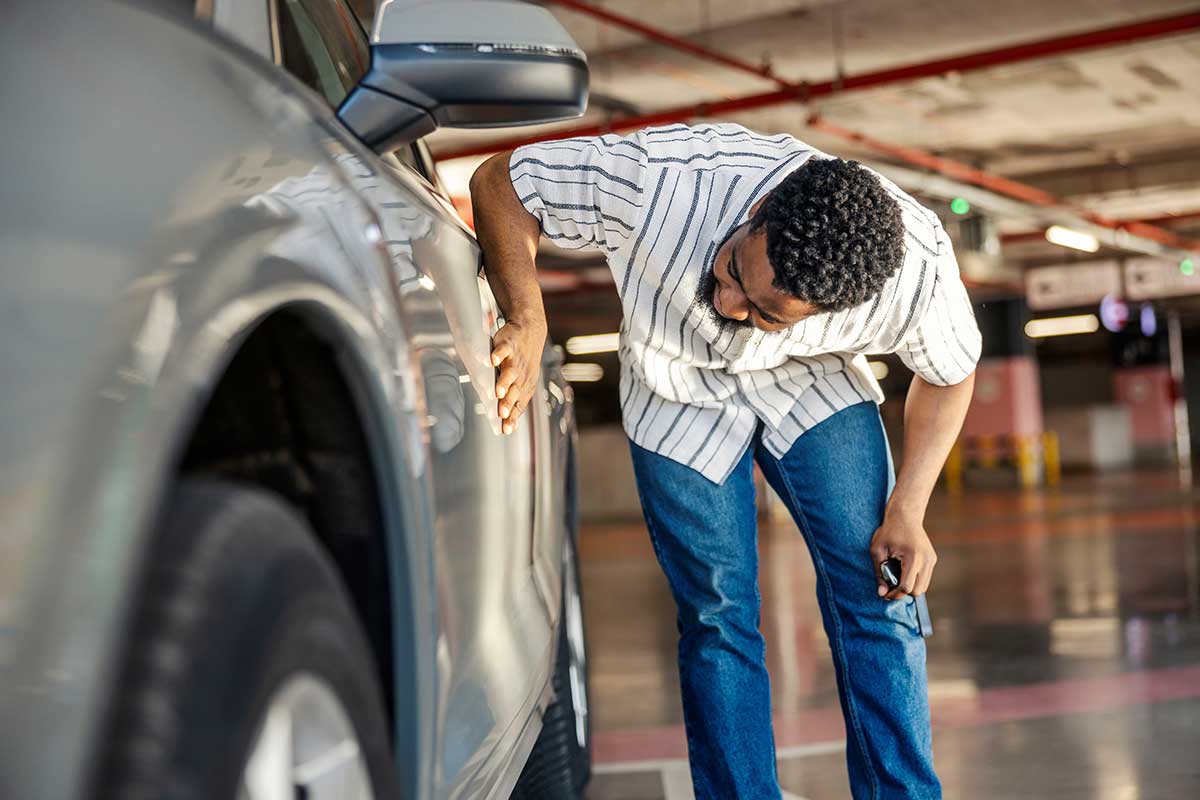 A man wearing a striped shirt and jeans leans over to closely inspect the side of a silver car in a parking lot.