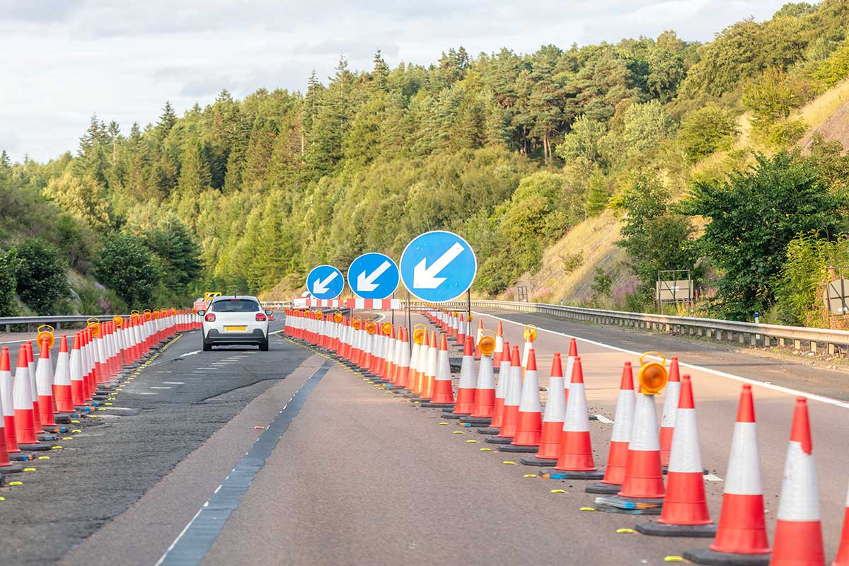 A white ATV drives along a road under construction, guided by lines of traffic cones and circular blue signs with white downward-pointing arrows.