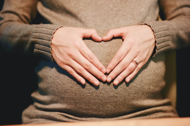 A pregnant person’s hands formed into a heart shape over their belly.
