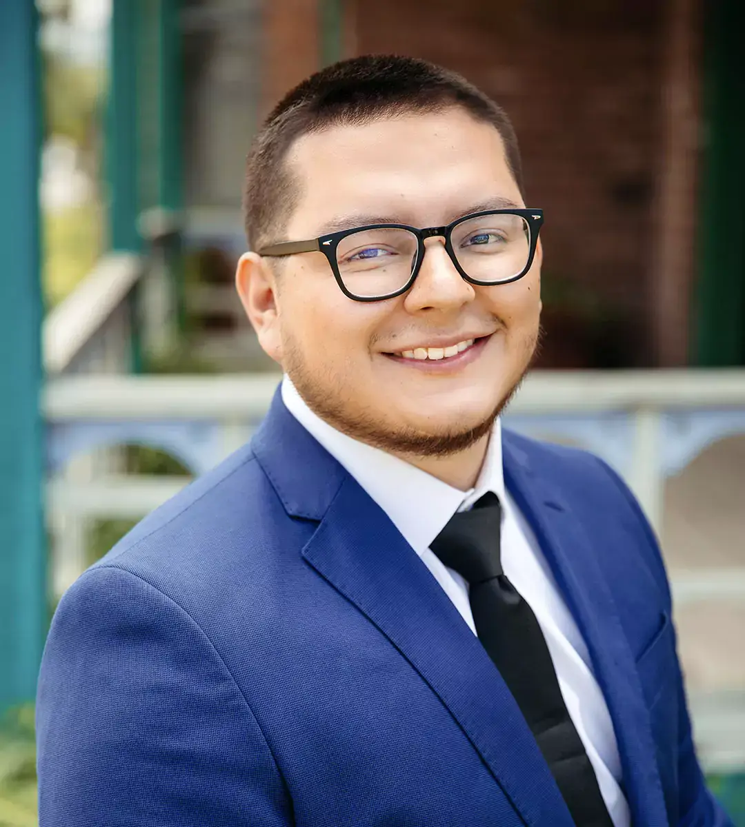 Rolando Meza is a young man with short dark hair and black-rimmed glasses, wearing a blue suit, white shirt, and black tie.