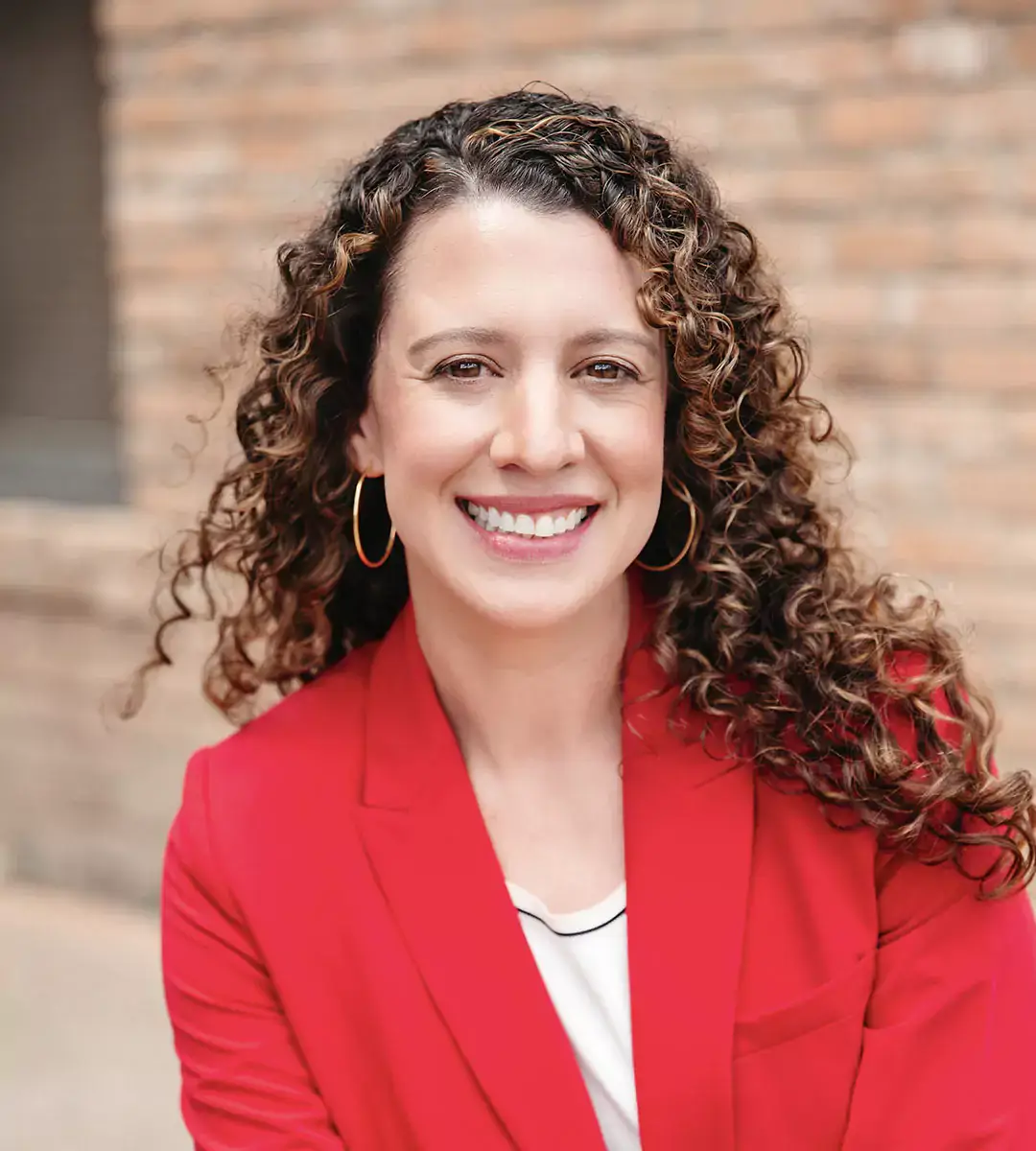 Valerie Cochlin smiles while wearing a red blazer and gold hoop earrings with her long curly hair.